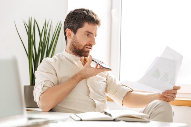 Imagen del empresario europeo de 30 años con camisa blanca hablando por teléfono móvil y sosteniendo un documento en papel, mientras trabaja en la oficina