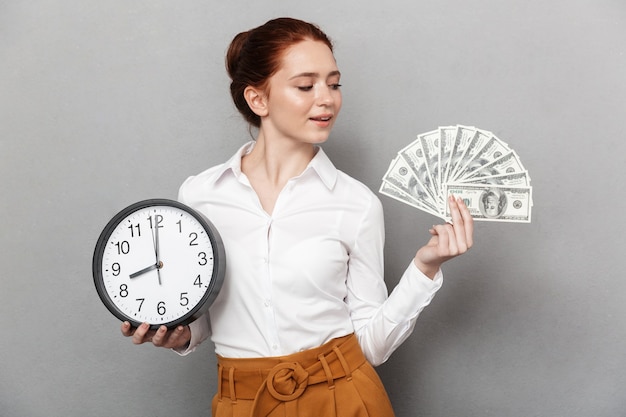 Imagen de la encantadora empresaria pelirroja de 20 años en ropa formal sosteniendo un gran reloj y un montón de dinero en efectivo aislado sobre gris