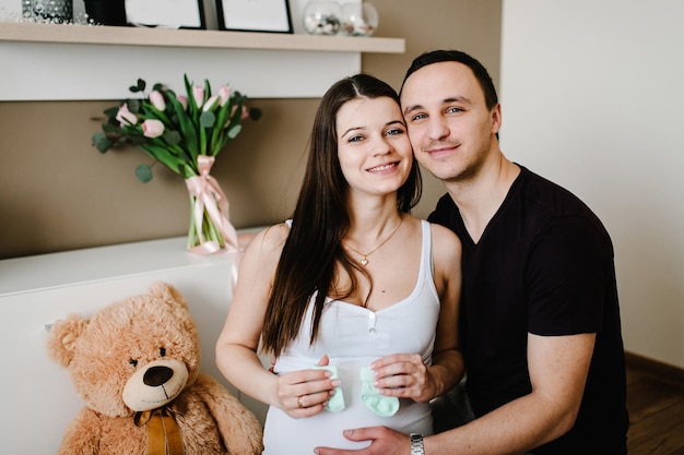 Imagen del esposo sosteniendo el vientre de su esposa embarazada calcetines para el bebé en las manos Mujer embarazada o niña y hombre guapo amoroso abrazando la barriga en casa Concepto de paternidad de pareja amorosa Baby Shower