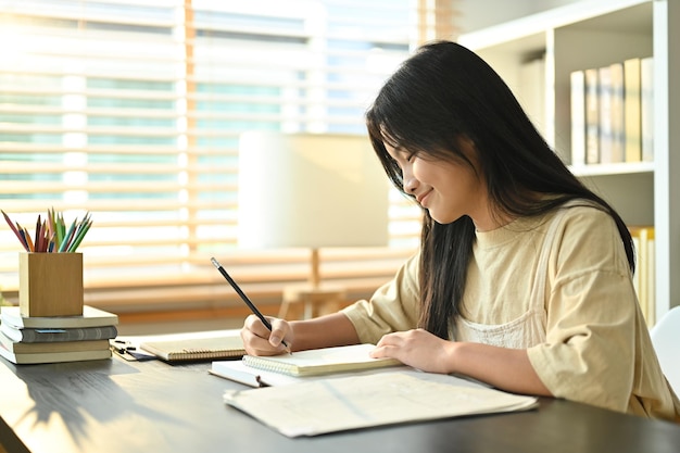 Imagen de un estudiante adolescente asiático sonriente haciendo tareas de escritura tomando notas en el escritorio en casa