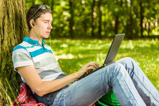 Imagen de estudiante con portátil y auriculares sentado bajo un árbol en el césped de verano