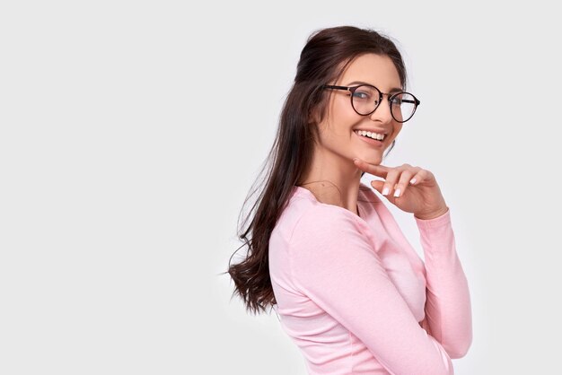 Imagen de estudio aislado de una mujer bastante joven con una amplia sonrisa que viste ropa informal y anteojos Mujer morena caucásica con blusa rosa posando sobre una pared blanca de estudio Concepto de personas y emociones