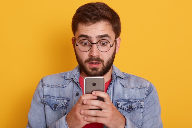 Imagen del excitado joven barbudo mirando el teléfono móvil con la boca abierta y la expresión facial sorprendida, posando en ropa elegante