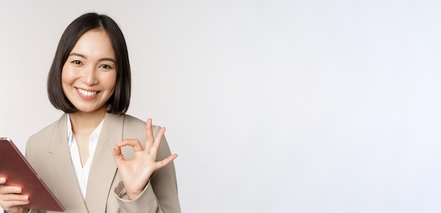 Imagen de una exitosa mujer de negocios asiática sosteniendo una tableta digital que muestra el signo de ok ok asegurando al cliente sobre fondo blanco