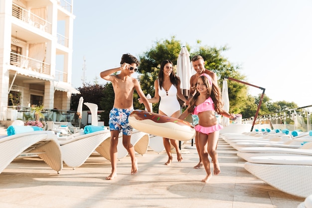 Imagen de familia caucásica feliz con niños descansando cerca de la piscina de lujo, con tumbonas y sombrillas de moda blanca fuera del hotel