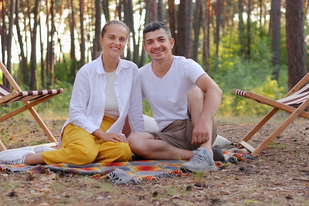 Imagen de una familia joven sonriente disfrutando del aire fresco y la hermosa naturaleza en el bosque sentada en una manta en el suelo mirando a la cámara feliz de pasar el fin de semana juntos