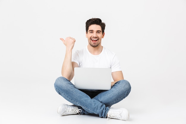 Imagen de feliz emocionado joven posando aislado sobre una pared blanca con ordenador portátil apuntando sentado en el suelo.