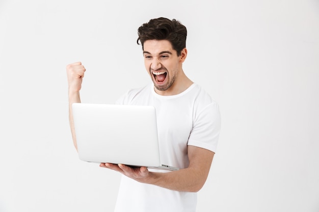 Imagen de feliz emocionado joven posando aislado sobre una pared blanca con ordenador portátil hacer gesto de ganador.