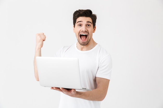 Imagen de feliz emocionado joven posando aislado sobre una pared blanca con ordenador portátil hacer gesto de ganador.