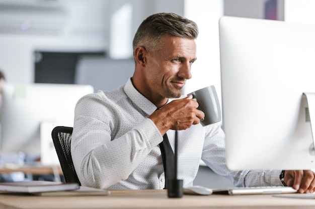 Imagen del feliz empresario de 30 años vistiendo camisa blanca y corbata bebiendo té de la taza, mientras está sentado junto a la computadora en la oficina