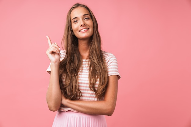 Imagen de feliz hermosa mujer vistiendo ropa a rayas sonriendo y apuntando con el dedo hacia arriba aislado sobre pared rosa