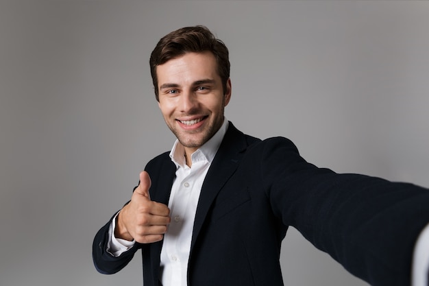 Imagen del feliz hombre de negocios de 30 años en traje formal mostrando el pulgar hacia arriba mientras toma una foto selfie, aislado sobre una pared gris