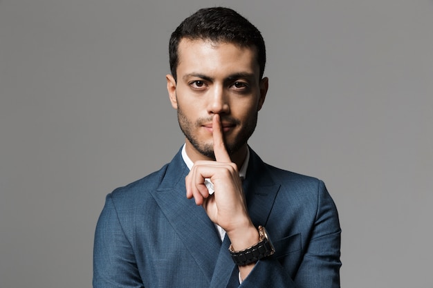 Imagen del feliz hombre de negocios árabe de 30 años en traje formal sosteniendo el dedo índice en los labios, aislado sobre pared gris