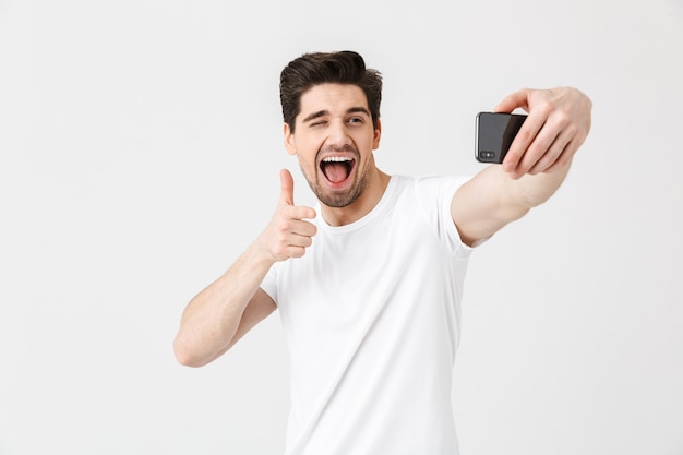 Imagen de feliz joven emocional posando aislado sobre pared blanca apuntando a tomar selfie por teléfono móvil.