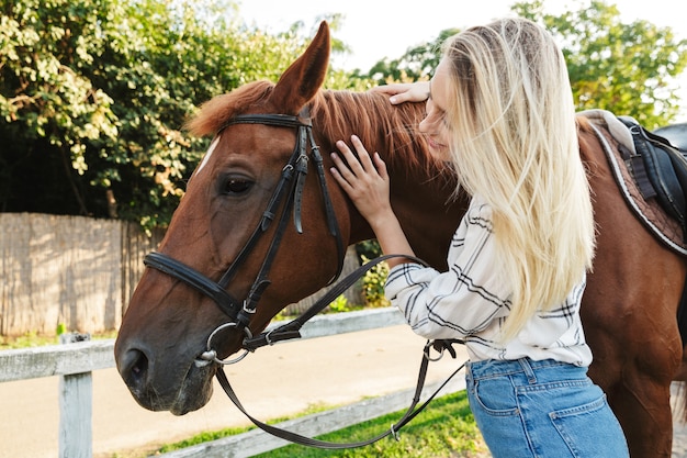 Imagen de feliz joven mujer rubia sonriendo y de pie a caballo en el patio en el campo