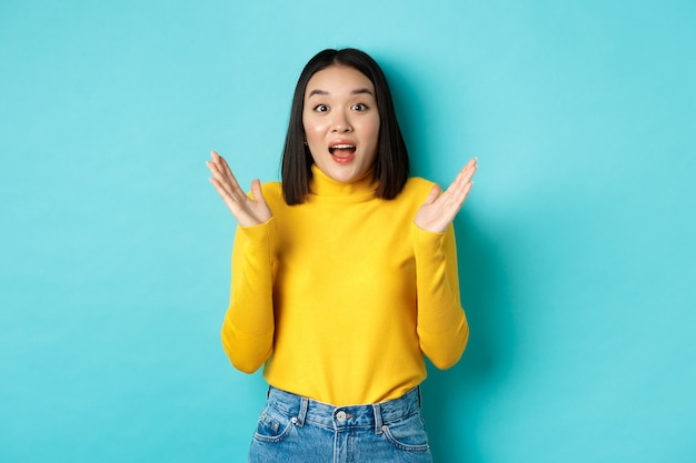 Imagen de feliz mujer asiática aplaudir y mirando asombrado, de pie en jersey amarillo sobre azul.