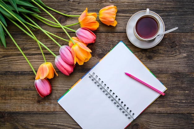 Imagen festiva romántica con hermosos tulipanes rosados y naranjas, una taza de té, un cuaderno sobre un fondo rústico de madera