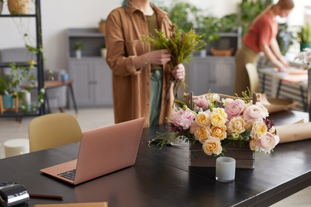 Imagen de fondo de la computadora portátil abierta en la mesa en la florería con floristería femenina irreconocible en el fondo, espacio de copia