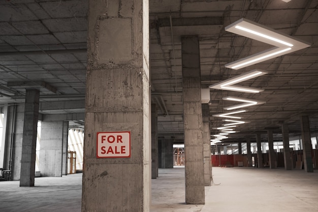 Imagen de fondo del edificio vacío en construcción con cartel de venta en columna de hormigón y lámparas de techo gráficas,