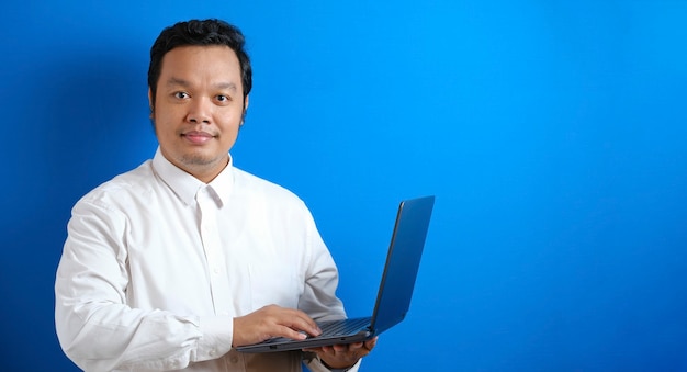 Imagen de la foto retrato de un hombre de negocios asiático adulto de pie y sonriendo mientras sostiene la computadora portátil y escribe sobre ella contra el fondo azul