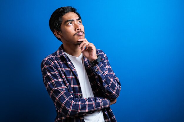 La imagen fotográfica de un joven estudiante asiático con camisa parecía feliz pensando y mirando hacia arriba, teniendo una buena idea. Retrato de medio cuerpo contra el fondo azul con espacio de copia