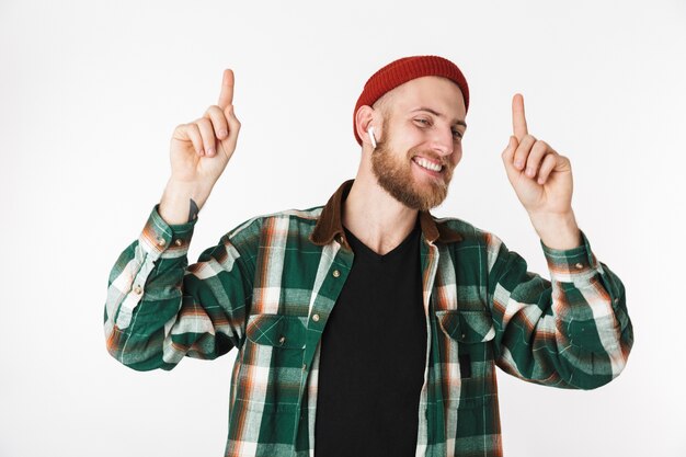 Imagen de guapo chico barbudo con sombrero y camisa a cuadros sonriendo, mientras está de pie aislado sobre fondo blanco.