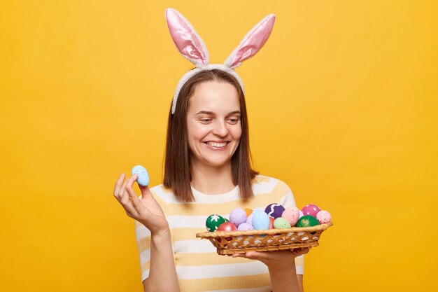 Imagen de una hermosa y alegre mujer europea sonriente con orejas de conejo sosteniendo huevos de Pascua mirando hacia otro lado disfrutando de la celebración de Pascua posando aislada en un fondo amarillo