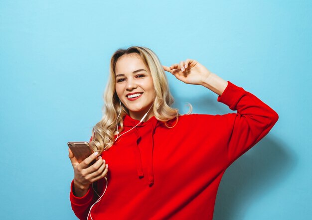 Imagen de una hermosa chica rubia excitada escuchando música en auriculares y bailando sobre la pared azul