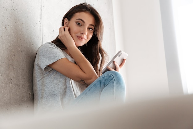 Foto imagen de la hermosa joven morena sonriendo y sosteniendo el teléfono inteligente sobre un muro de hormigón en casa