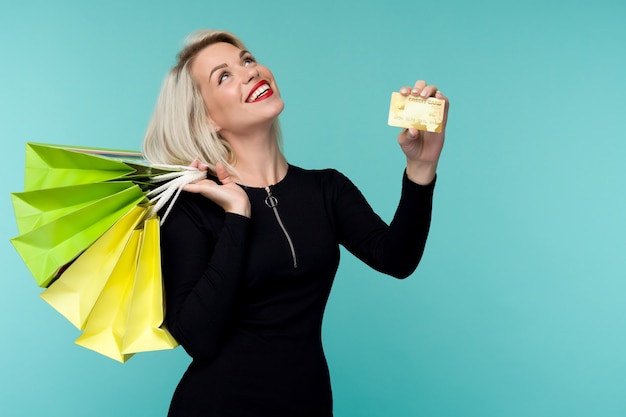 Imagen de una hermosa joven rubia feliz posando aislado sobre fondo de pared azul sosteniendo bolsas de la compra.