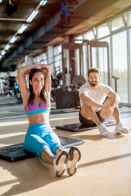 Imagen de la hermosa joven sonriente deportiva estirando los músculos de su brazo después del entrenamiento mientras su entrenador personal la está mirando.