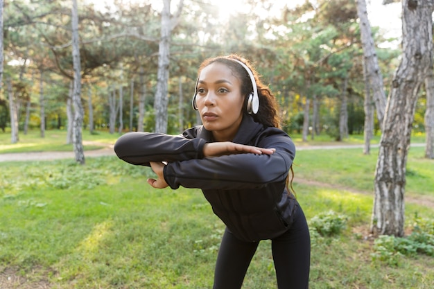 Imagen de hermosa mujer de 20 años vistiendo chándal negro haciendo ejercicio y estirando el cuerpo en el parque verde