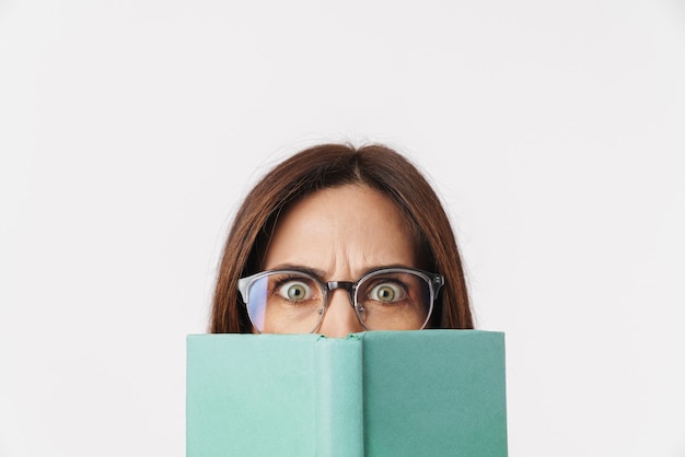 Imagen de hermosa mujer adulta morena con anteojos frunciendo el ceño y coning su rostro con libro aislado en blanco