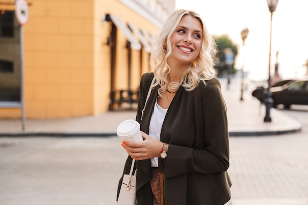 Imagen de hermosa mujer con chaqueta sosteniendo café para llevar en vaso de papel, mientras camina por las calles de la ciudad