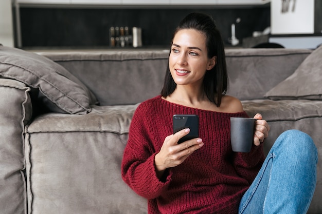 Imagen de una hermosa mujer en el interior de la casa cerca del sofá con teléfono móvil tomando café.