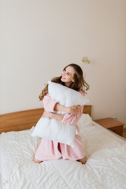 Imagen de hermosa mujer joven increíble en el interior posando en casa con una túnica blanca cerca de grandes ventanales.