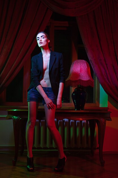 La imagen de una hermosa mujer de moda de pie junto a la mesa con lámpara frente a la ventana con cortina