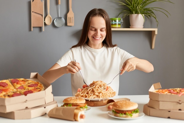 Imagen de una hermosa mujer optimista con el pelo castaño y una camiseta blanca informal sentada en la mesa en la cocina comiendo pasta con tenedores sonriendo con la cara satisfecha disfrutando de la comida chatarra