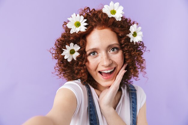 Imagen de una hermosa pelirroja feliz rizada posando aislada sobre pared púrpura con flores en el cabello tomar selfie.