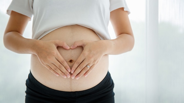 Una imagen de hermosas manos con una bonita piel clara haciendo un gesto de forma de corazón en el vientre del embarazo sobre fondo blanco. Concepto de madre sana y feliz.