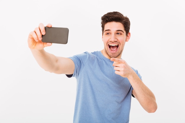 Imagen del hombre 30s con cabello castaño sonriendo y señalando con el dedo a la cámara mientras toma selfie en teléfono celular negro, aislado sobre la pared blanca