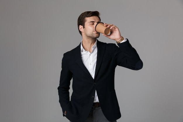 Imagen de un hombre adulto de 30 años en traje formal bebiendo café de un vaso de papel, aislado sobre una pared gris