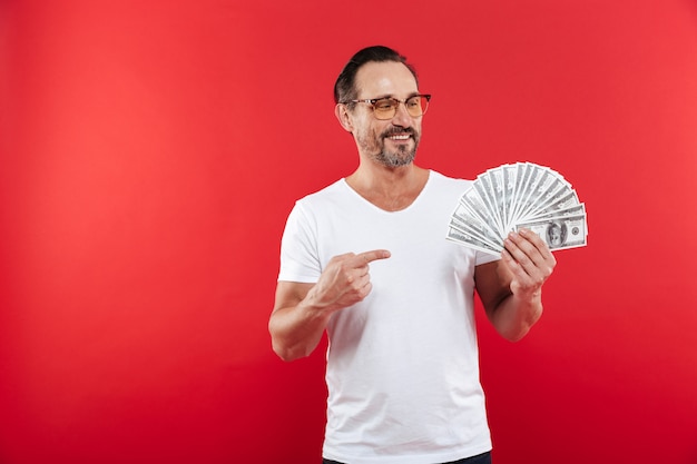 Imagen de hombre adulto 30s en camiseta blanca casual con gafas sonriendo y señalando con el dedo en un montón de dinero en efectivo en dólares en moneda en la mano, aislado sobre fondo rojo.