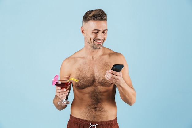 Imagen de un hombre adulto feliz emocionado guapo posando sobre pared azul bebiendo cócteles mediante teléfono móvil.