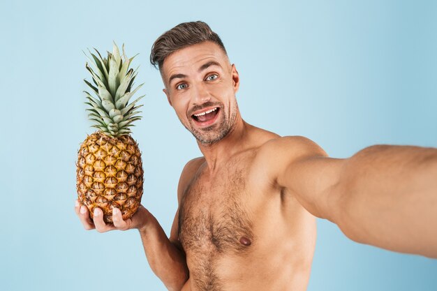 Imagen de un hombre adulto feliz emocionado guapo en traje de baño posando sobre pared azul tomar un selfie por cámara con piña.