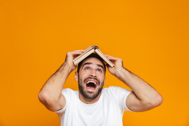 Imagen de hombre alegre de 30 años en camiseta blanca sosteniendo un libro sobre su cabeza, aislado