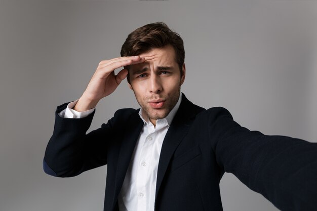 Imagen de hombre alegre de 30 años en traje formal manteniendo la mano en la frente mientras toma una foto selfie, aislado sobre una pared gris