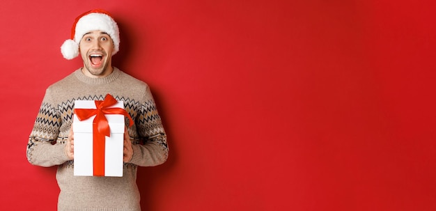 Imagen de un hombre apuesto y emocionado que recibe un regalo de Navidad, usa un sombrero de Papá Noel y un suéter de invierno, grita de alegría, sostiene un regalo y se para sobre un fondo rojo.