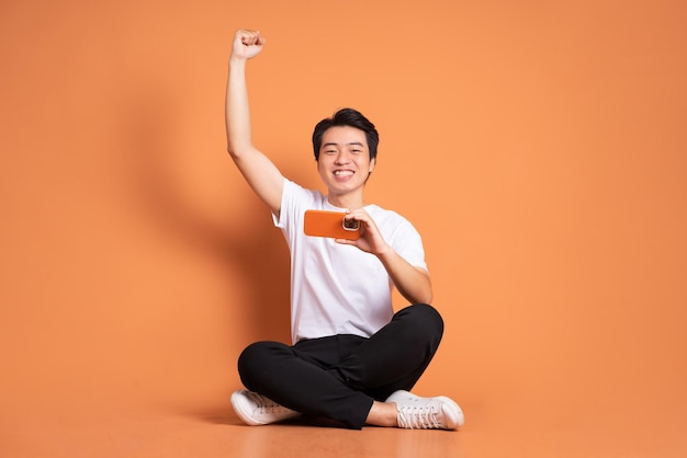 Imagen de un hombre asiático sentado usando un teléfono y aislado en un fondo naranja