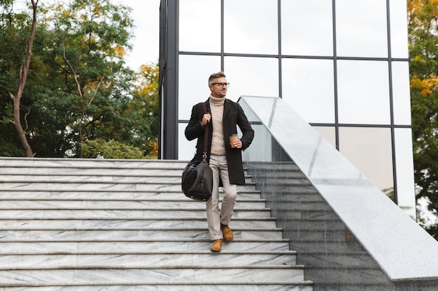 Foto imagen de hombre barbudo de 30 años con anteojos, caminando por las calles de la ciudad y sosteniendo café para llevar